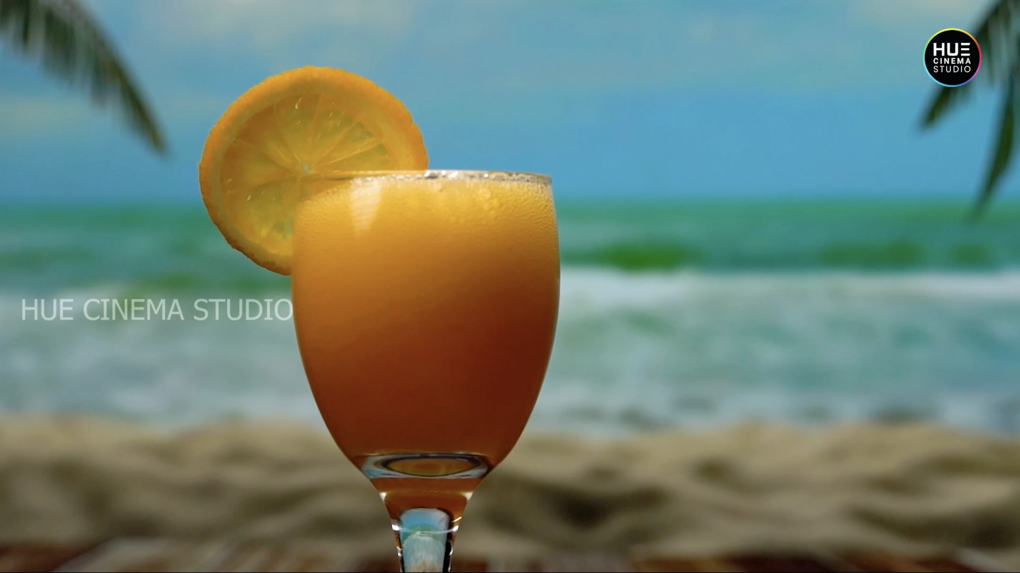 A tulip glass of orange juice garnished with an orange slice against a summer beach backdrop, from the Alkor Cyber Space juices b-roll showreel by Hue Cinema Studio.